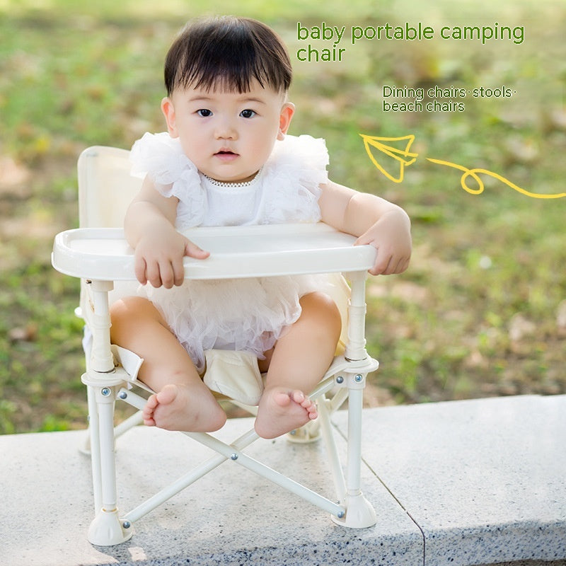 A baby in a white fluffy outfit sits on a portable camping chair with a tray, outdoors on a stone surface. Green grass is visible in the background.