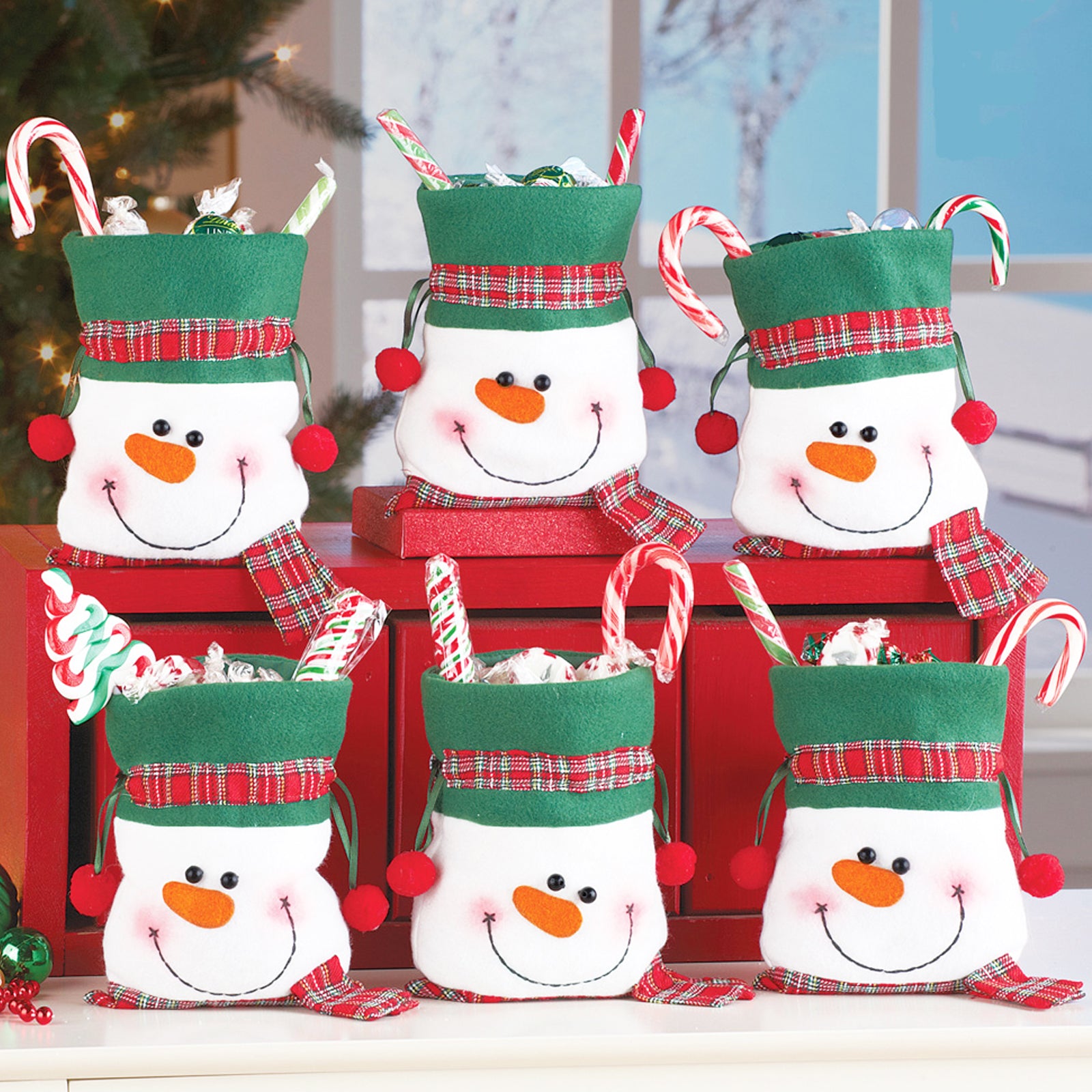Six snowman-themed gift bags with candy canes, wrapped treats, and festive decorations. They feature green hats, plaid accents, and smiling faces, set on a red surface.