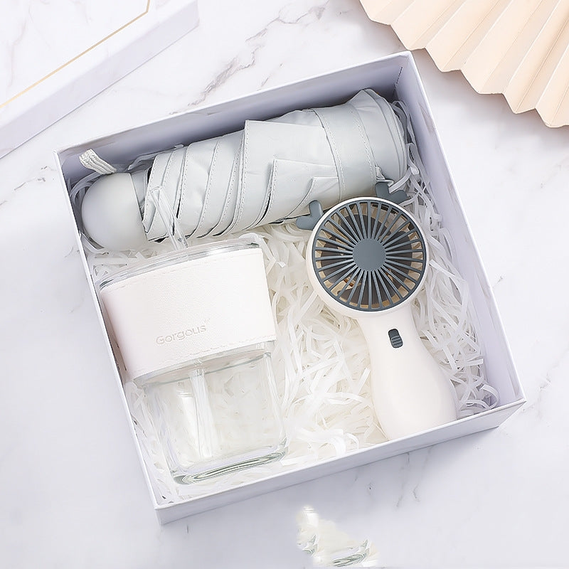 A gift box containing a folded gray umbrella, a portable white fan, and a clear white-capped bottle, all nestled in white shredded paper on a marble surface.