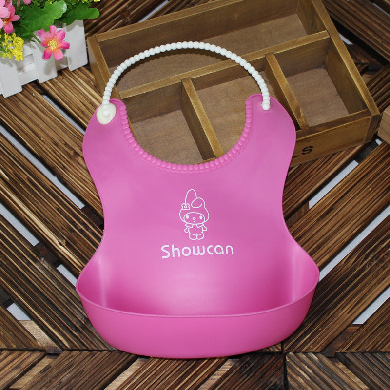 Pink silicone baby bib with cartoon character design and white pearl necklace strap, placed on a wooden surface next to a wooden box and flowers.