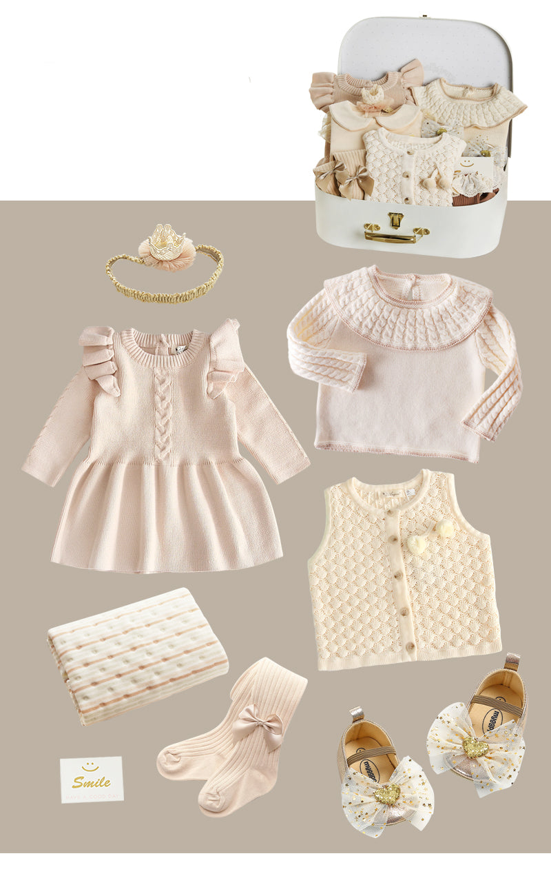 Baby clothing set including a pink dress, sweater, ivory vest, socks, shoes with bows, a blanket, and a headband, all neatly arranged alongside a white suitcase.