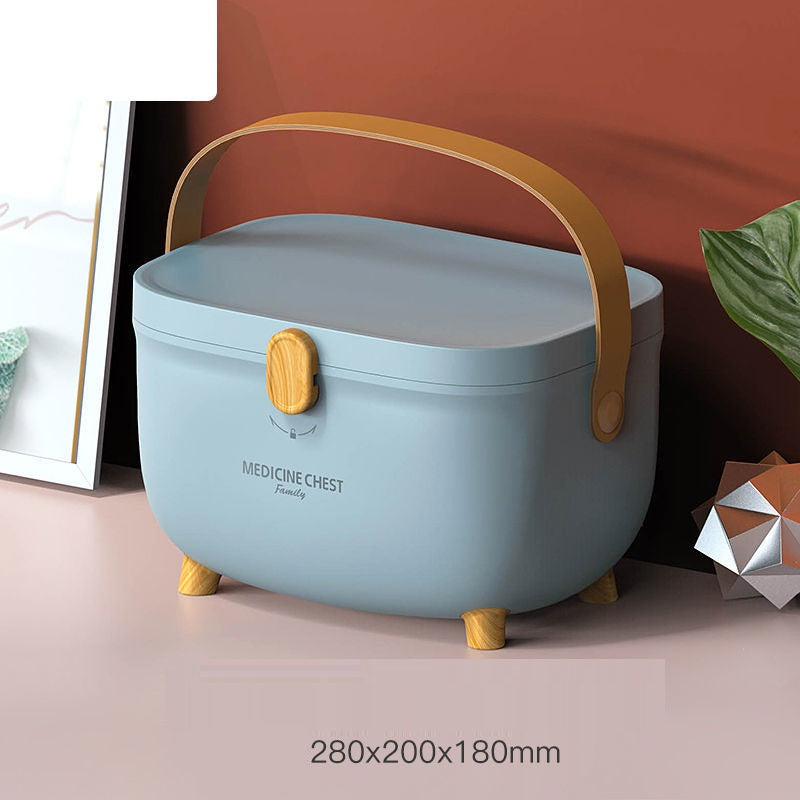 A light blue medicine chest with a tan handle and latch, resting on small wooden legs. It is placed on a pink surface against a reddish-brown wall, with visible dimensions 280x200x180mm.