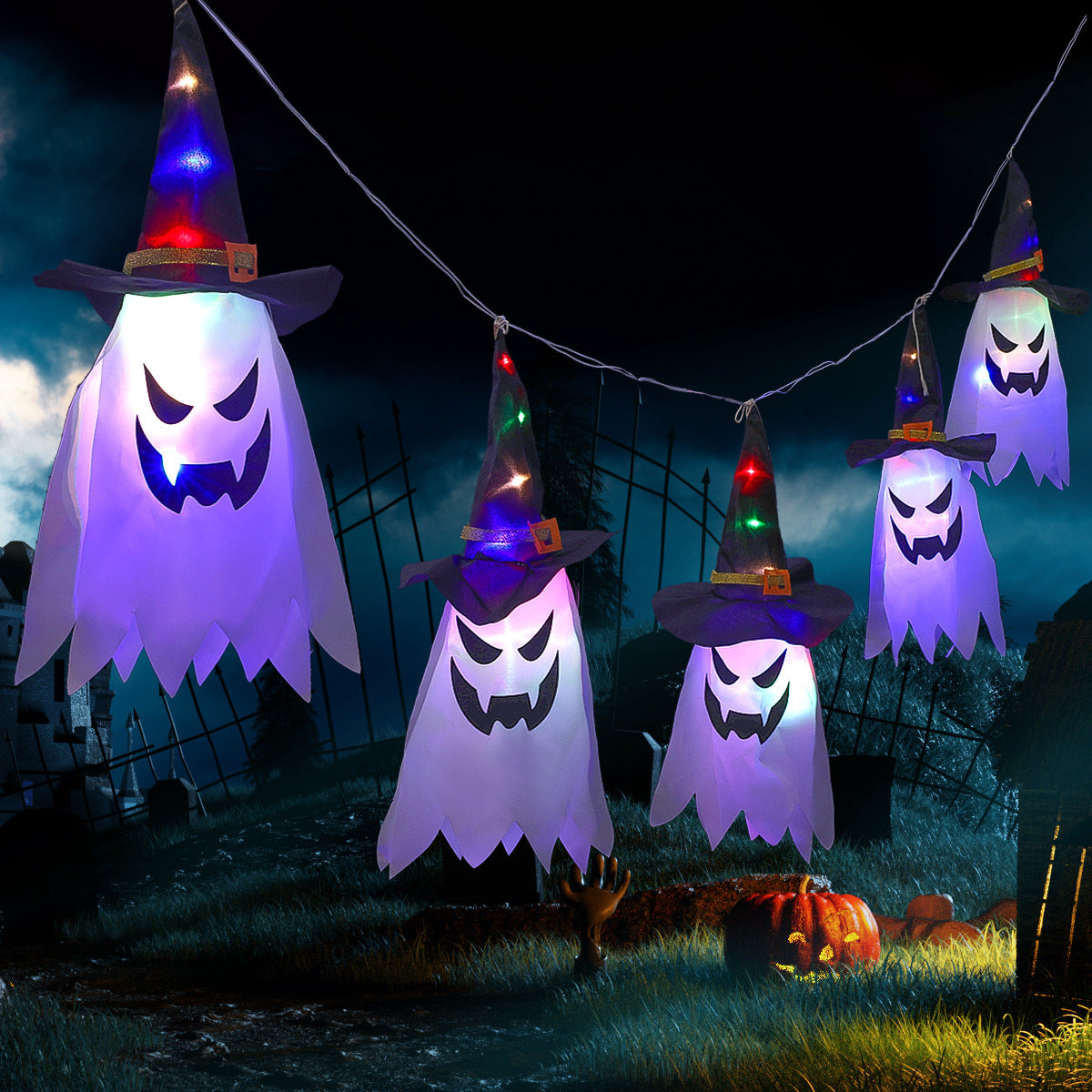 String of glowing ghost decorations with witch hats, featuring eerie faces, hang in a dark, spooky outdoor setting with a jack-o'-lantern and a skeletal hand emerging from the ground.