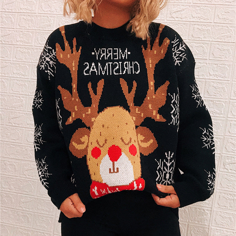 Person wearing a black sweater featuring a reindeer with a red nose, surrounded by snowflakes, and backward text "Merry Christmas." The background is a white textured wall.