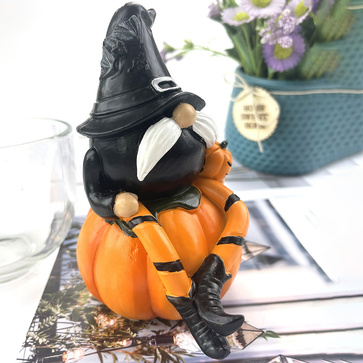 A whimsical gnome with a black hat and white mustache sits on an orange pumpkin. The gnome wears striped leggings and boots. A vase with purple flowers is in the background.