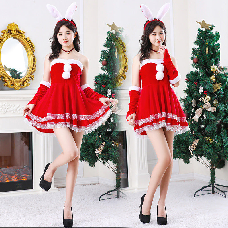 Two women in red Santa-style dresses with white trim and bunny ears pose playfully by decorated Christmas trees, with a fireplace and ornate mirror in the background.