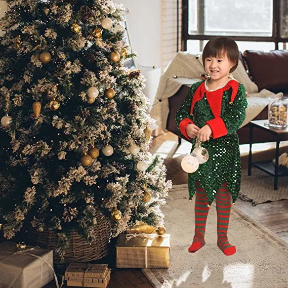 A child in a sequined green elf costume stands next to a decorated Christmas tree with gold and white ornaments, holding a gift, in a cozy living room with wrapped presents.