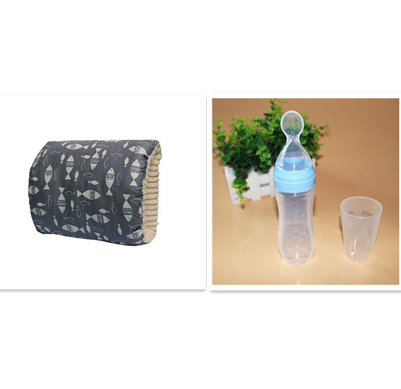 A baby bottle with a blue cap and spoon design sits beside a transparent cup, with a small plant in the background. Next to it is a dark pillow with white fish patterns.