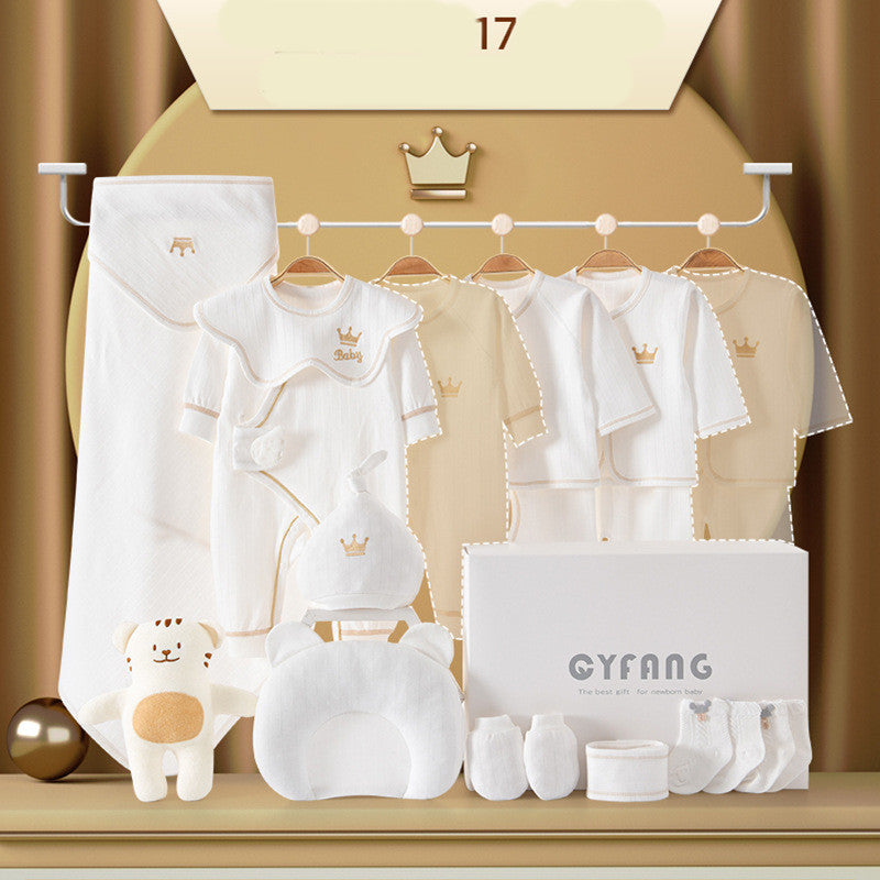 A display of baby clothing and accessories, including white and beige outfits with crown motifs, a plush toy, a pillow, socks, and a gift box, set against a soft backdrop.