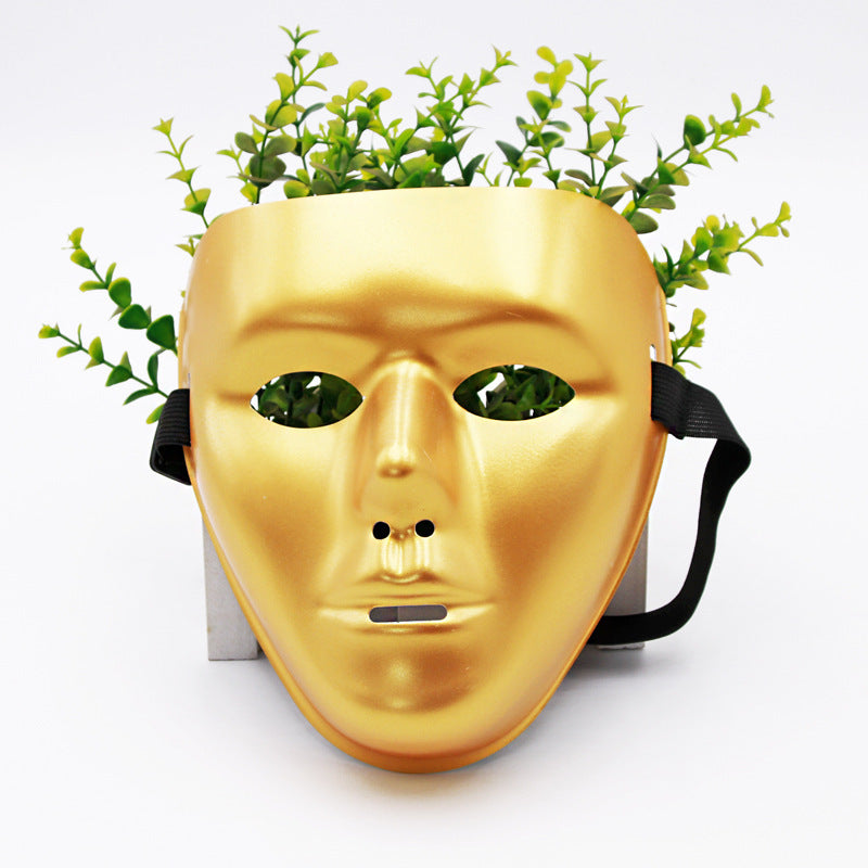 A golden mask with eye holes and straps is placed in front of green leafy plants, giving the appearance of the mask wearing the plants like hair.