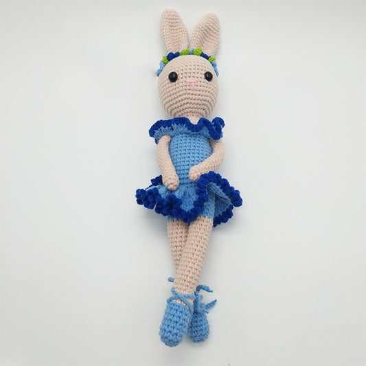 Crocheted bunny doll in a blue dress with ruffled edges, wearing blue shoes and a colorful flower headband, sits with legs folded on a white background.