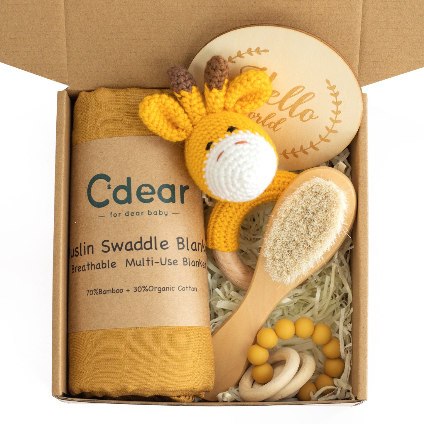 Gift box with a mustard muslin swaddle blanket, a knitted giraffe rattle, a wooden baby brush, a beaded teether, and a "Hello World" wooden plaque on shredded paper.