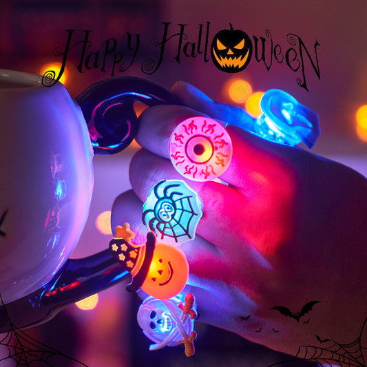 Fingers with glowing Halloween rings featuring a spider, pumpkin, eye, and ghost hold a cup with “Happy Halloween” text above. Background has blurred orange lights.