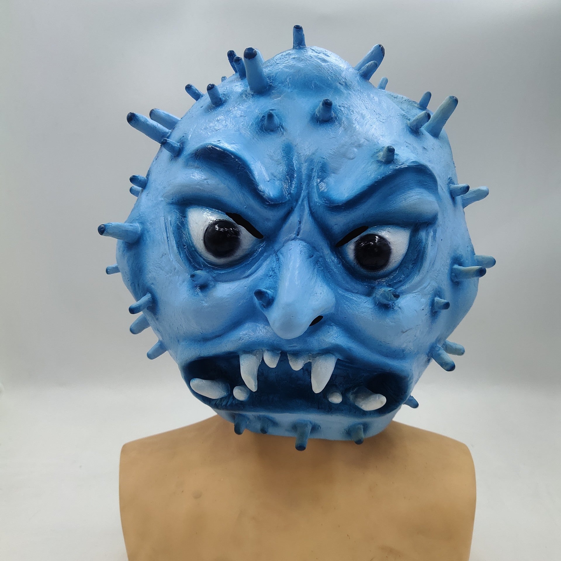Blue mask shaped like a virus with angry eyes, sharp teeth, and spikes. The mask has an expressive, intense look and is mounted on a plain mannequin bust.