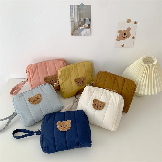 Six quilted pouches in pastel shades with bear patches arranged on a table. A pleated lamp and wall decorations, including photos and a bear illustration, are in the background.