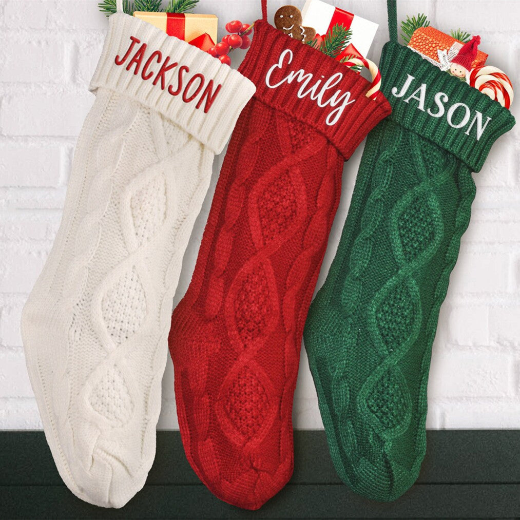 Three knitted Christmas stockings hang against a white brick wall, labeled with names. They are cream, red, and green, filled with gifts and festive items like candy canes.