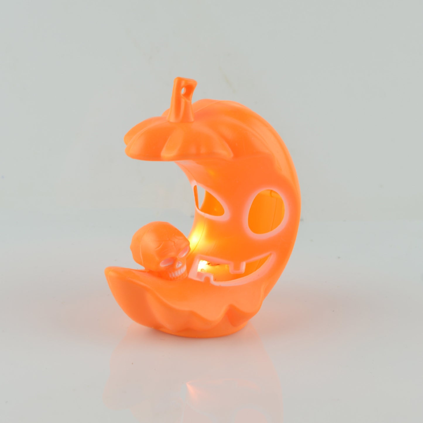 Glowing orange crescent-shaped pumpkin decoration with carved face, featuring a small skull inside, on a reflective white surface.