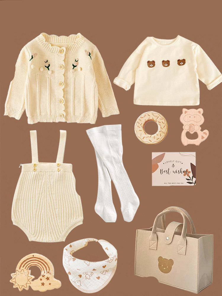 Baby gift set with a knitted cardigan, bear-embroidered top, suspenders, tights, teething rings, bib, a greeting card, and a bag with bear face design, all in neutrals.