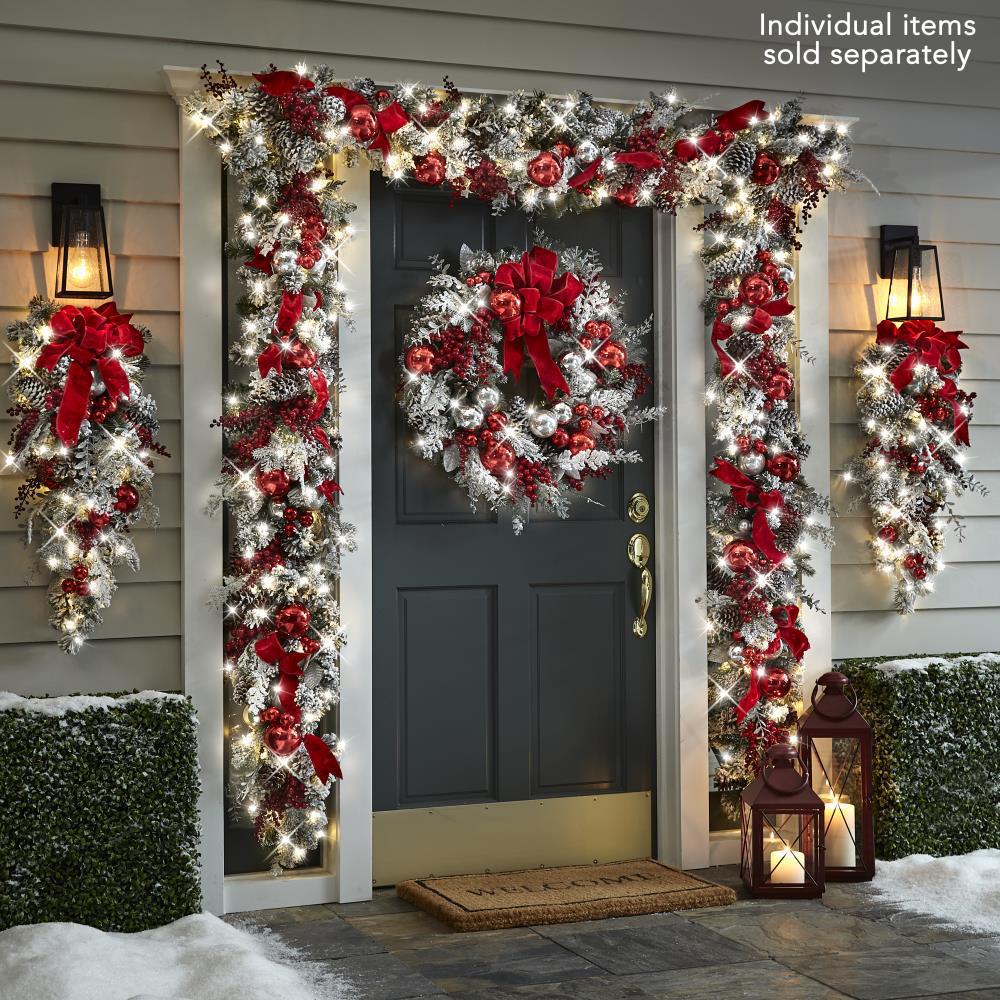 Festive front door decorated with red and silver garland, large wreath with bows, and bright lights. Two lanterns on the doorstep glow warmly against a snowy background.