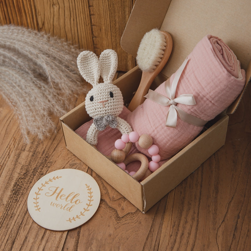 A gift box contains a pink blanket, a stuffed crocheted bunny, a wooden baby brush, and a teething ring. Nearby, a wooden "Hello world" sign sits on a wooden surface.