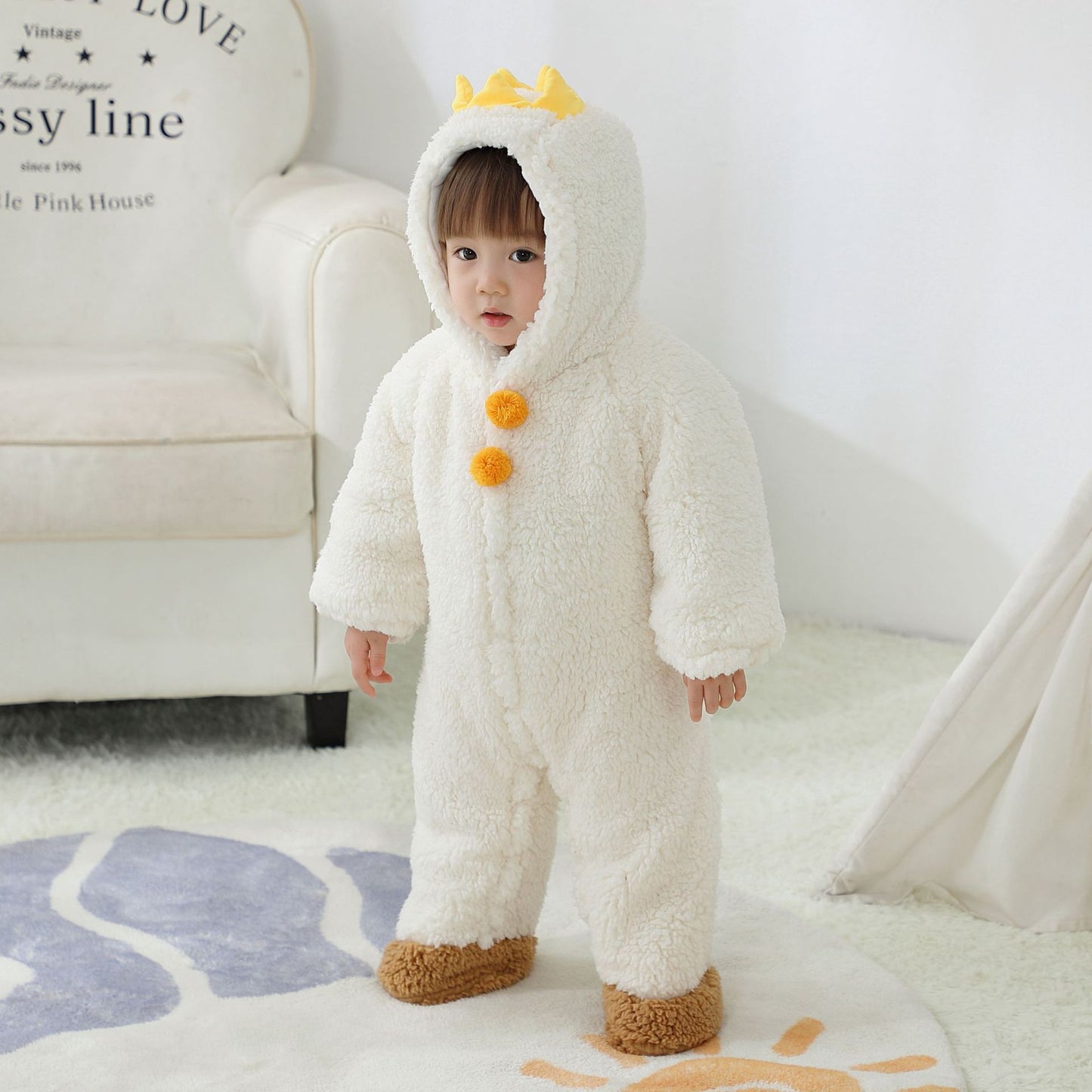 A toddler in a fluffy white onesie with yellow pom-poms and a crown on the hood stands on a patterned rug in a cozy room, near a white armchair and a beige tent.