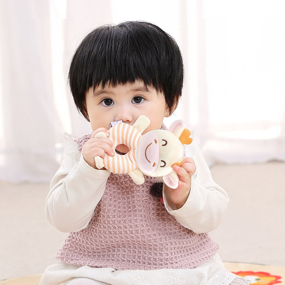 A young child with short black hair holds a plush toy with a cartoon face and striped body. The child wears a knitted pink vest over a white top, sitting indoors on a soft surface.