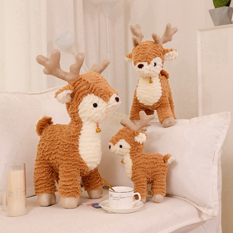 Three plush reindeer toys with brown fur and white bellies are displayed on a white couch. Each has antlers and wears a small bell collar. A teacup sits in front.