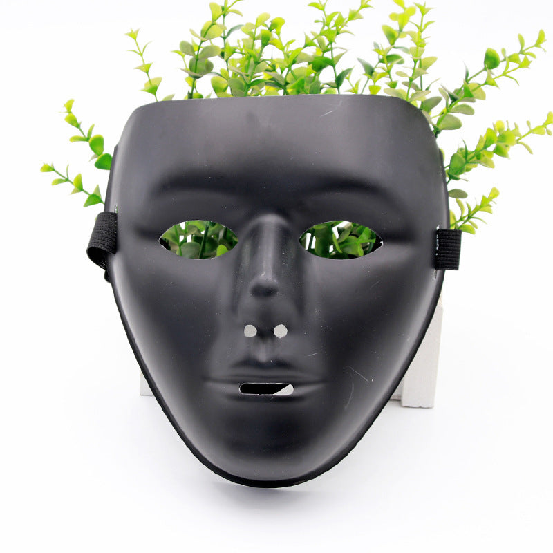 A black masquerade mask with cutouts for eyes and mouth, adorned on a white stand. Green foliage is visible behind the mask, resembling hair.