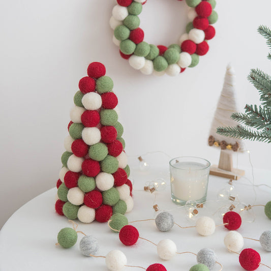 Felt ball tree and wreath in red, white, and green on a table, with a candle, string lights, and a mini yarn cone tree, creating a festive holiday scene.