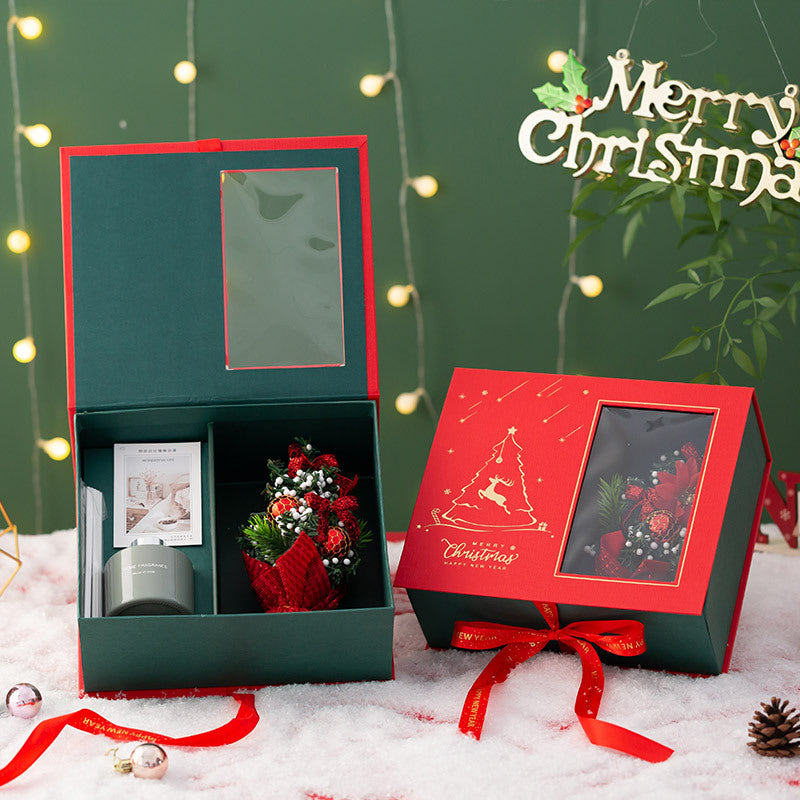 Christmas gift box set with festive design, featuring a candle, a small bouquet, and a card. The box is open, with a "Merry Christmas" sign in the background.