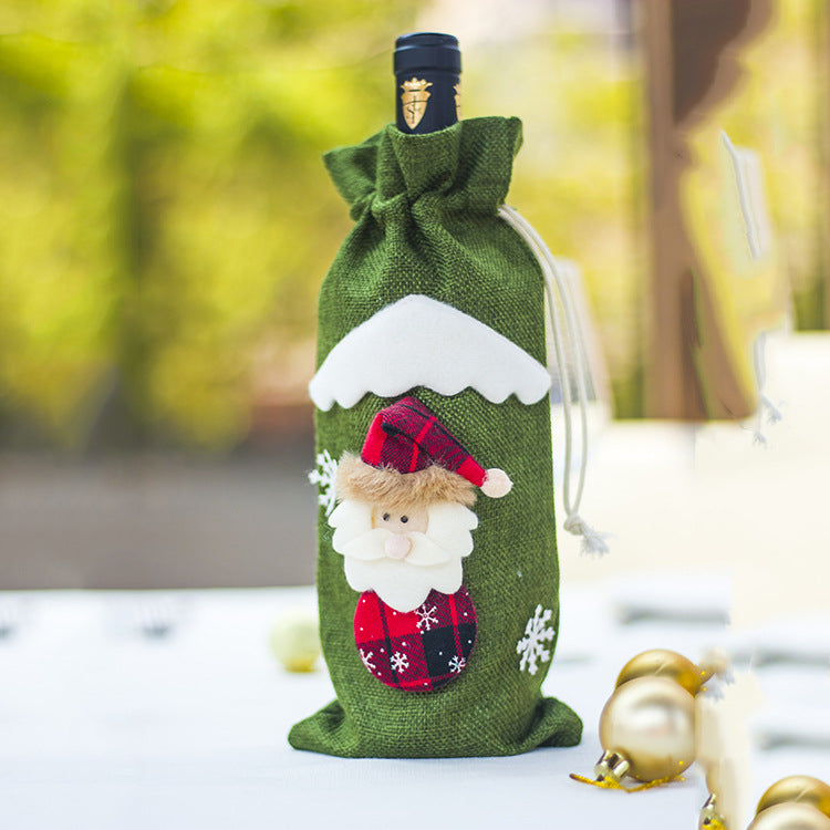 A wine bottle wrapped in a green festive cover featuring a Santa Claus design and snowflakes, with a snowy peak detail at the top. The background is softly blurred.