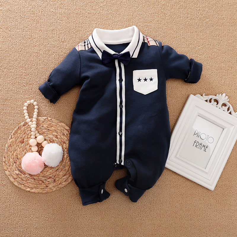 Navy blue baby onesie with white trim, a bow tie, and star pocket detail, laid on a woven mat. Beside it, a photo frame and a toy with pink and white pom-poms.