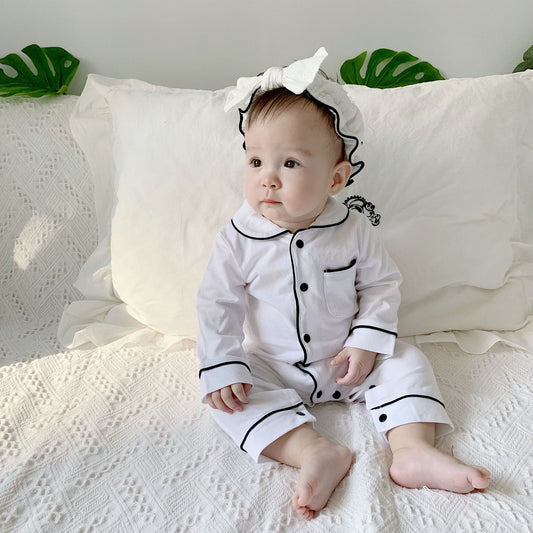 Baby in white pajamas with black trim and a large white headband sits on a textured white blanket. Green leaves decorate the background, creating a soft, cozy atmosphere.