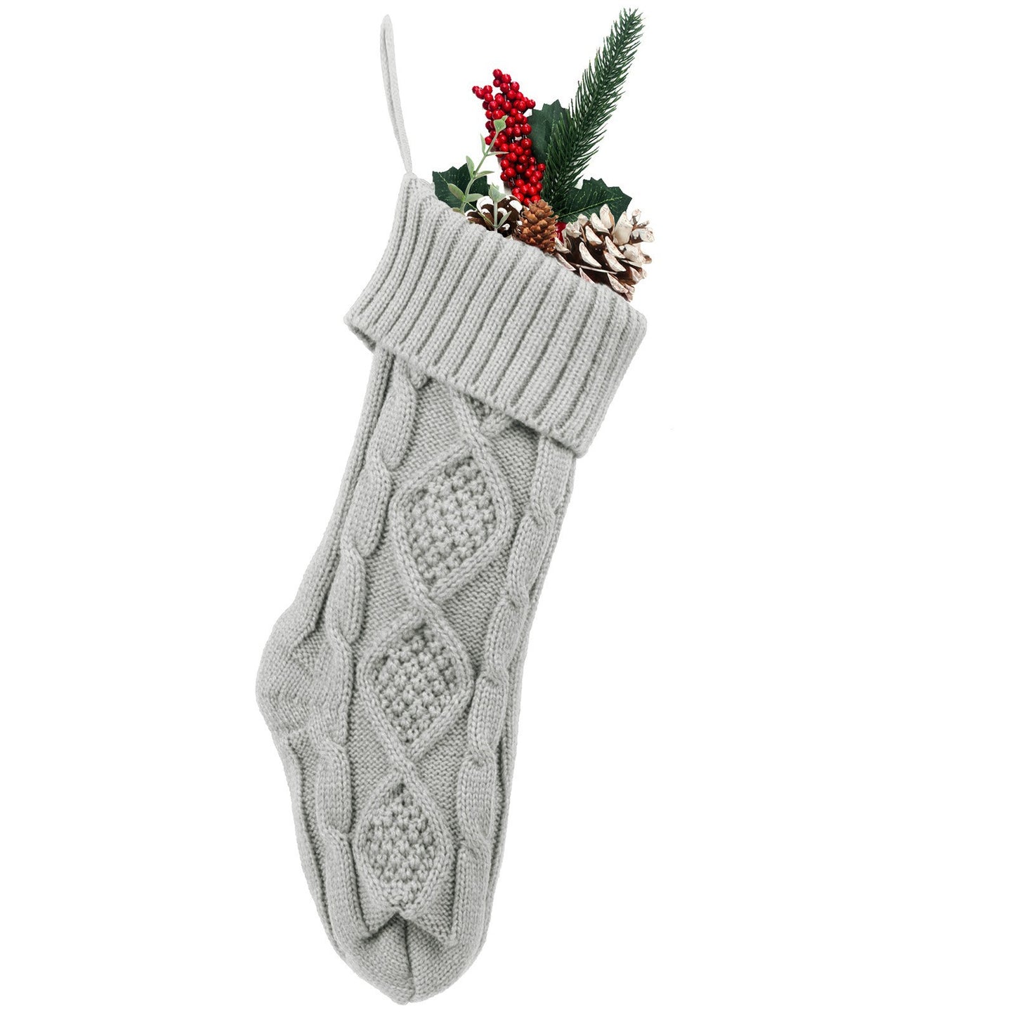 A gray knitted Christmas stocking filled with pinecones, red berries, holly leaves, and a pine branch, hanging against a white background.