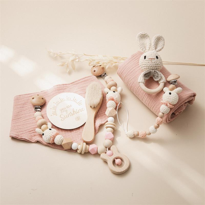 Baby accessories on a beige background, including a pink muslin cloth, a wooden brush, crochet bunny pacifier clip, wooden teether, and a "You are my Sunshine" card.
