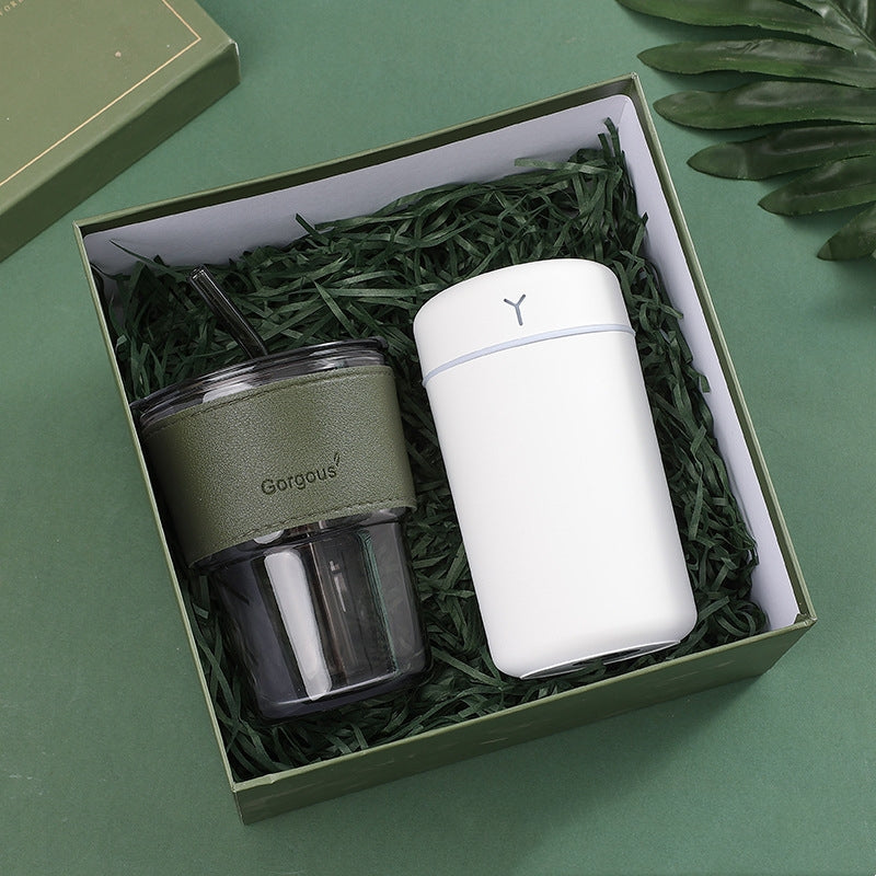 A gift box with a dark green insulated tumbler and a sleek white bottle, nestled in shredded green paper, set against a matching green backdrop with a leaf accent.
