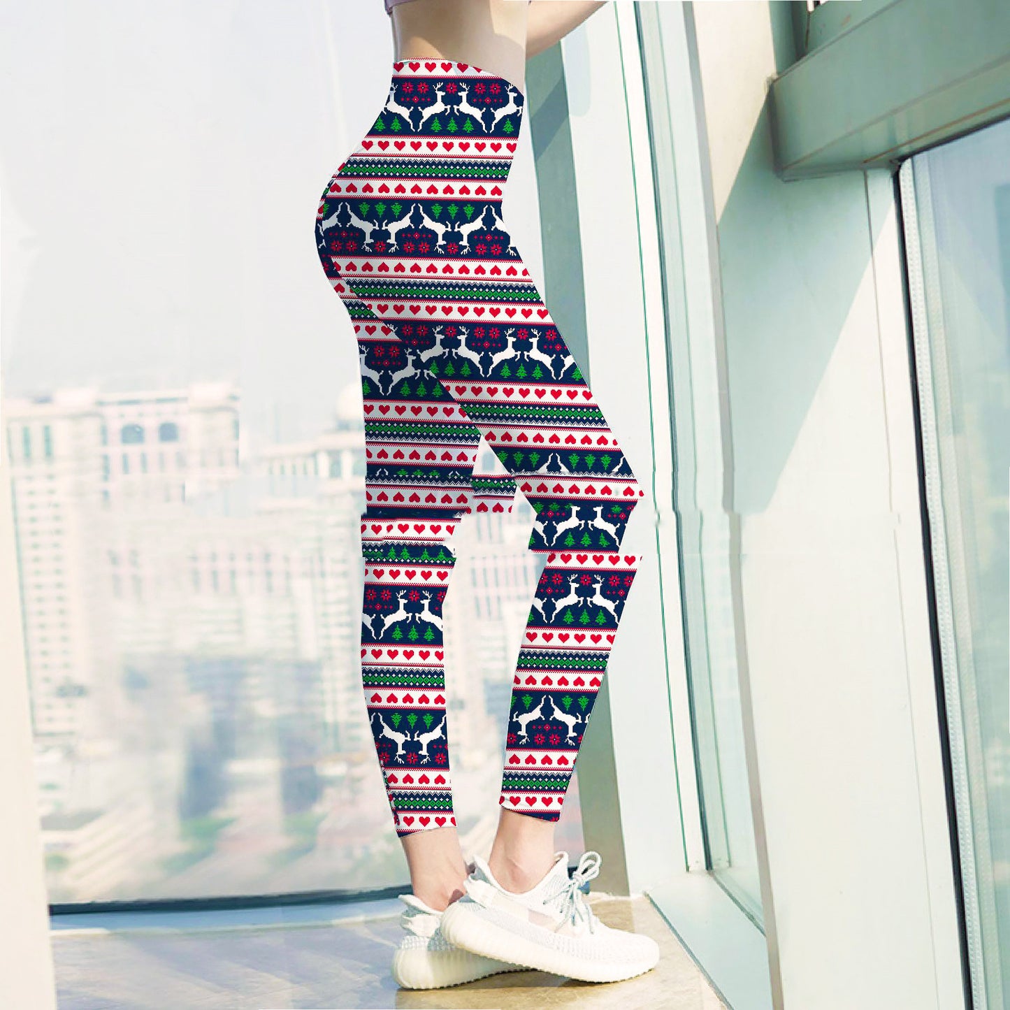 Person wearing festive leggings with reindeer and heart patterns, paired with white sneakers, stands by large windows with a blurred cityscape in the background.