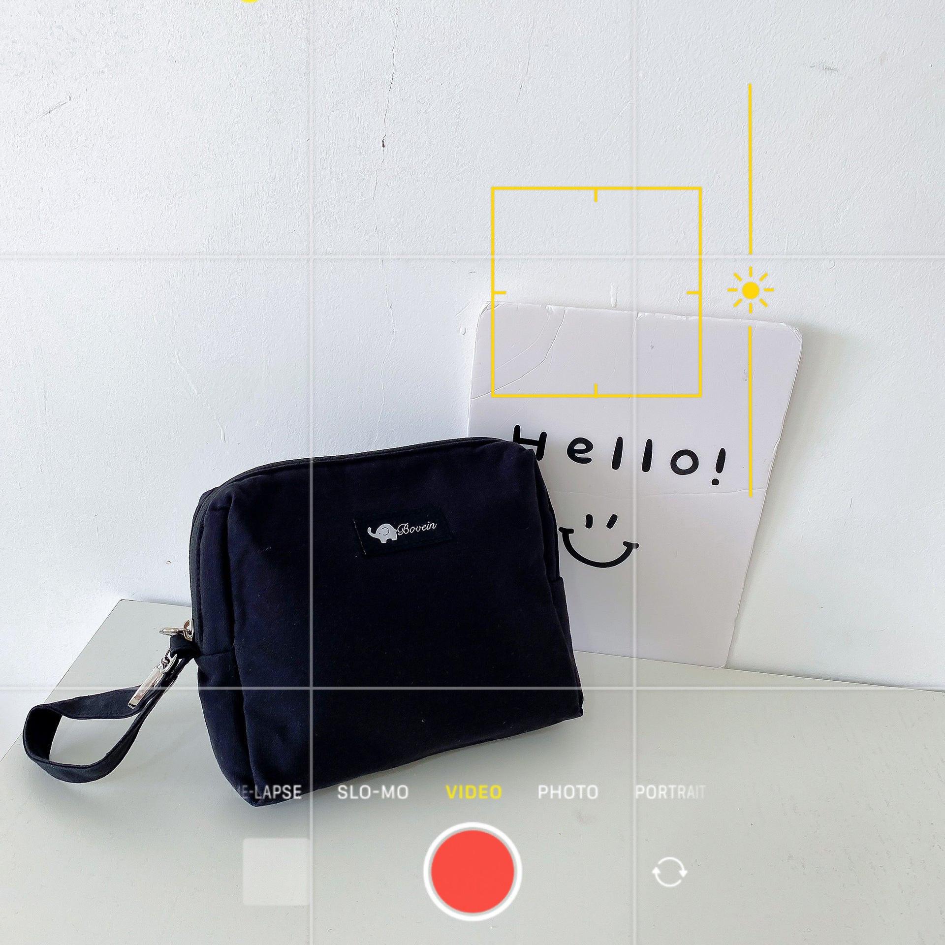 A black pouch on a white surface is next to a handwritten card saying "Hello!" with a smiley face. The camera interface is visible, highlighting "VIDEO" mode.