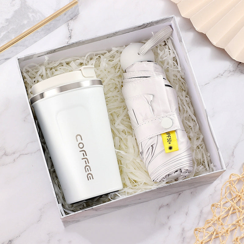 White coffee tumbler and folded umbrella in a gift box with white shredded paper, set on a marble surface. The umbrella tag reads "UPF 50+".