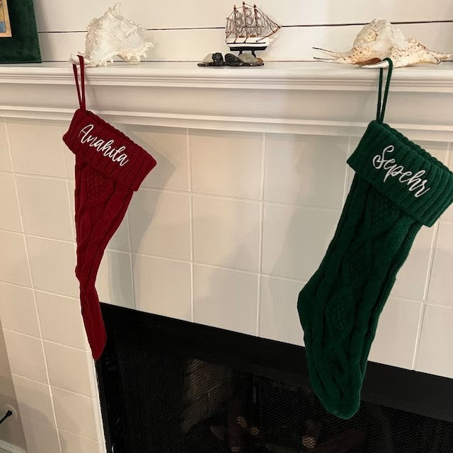 Two Christmas stockings hang on a white tiled fireplace. The red stocking is on the left and the green on the right. Seashells and a model ship decorate the mantel.