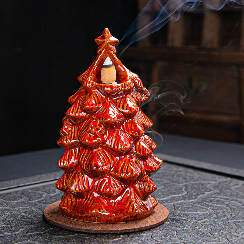 Red ceramic Christmas tree incense burner with smoke rising from the top, placed on a coaster against a dark background.