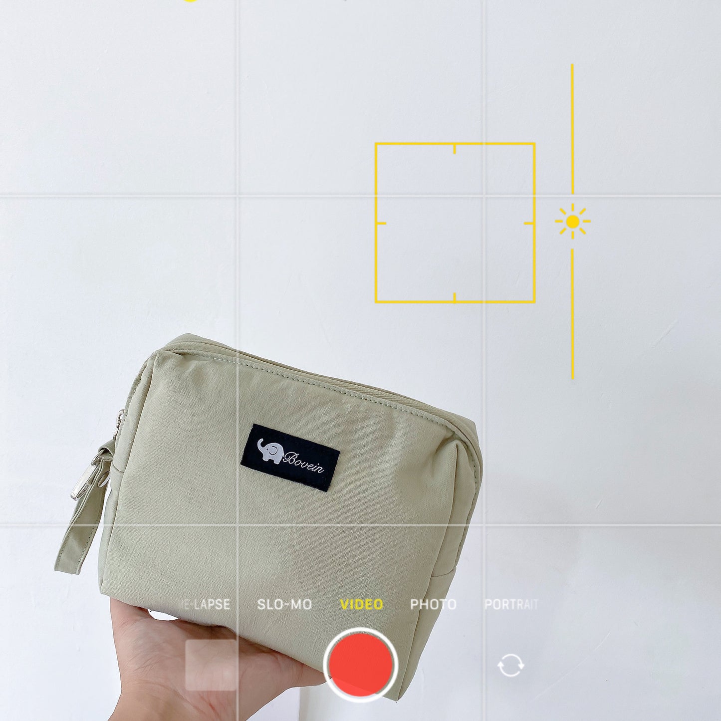 A hand holds a beige pouch with a black label showing an elephant logo and "Bovein" text. Camera interface overlays the image, showing video mode settings.