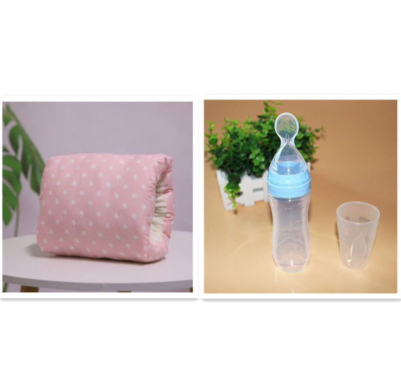 A soft pink baby blanket with white stars sits on a table, next to a baby bottle with a blue cap and clear spoon, accompanied by a small clear cup, with greenery in the background.