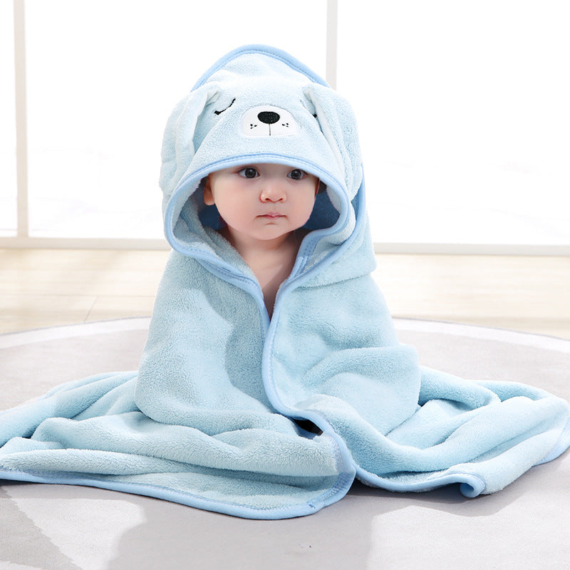 A baby wrapped in a soft blue blanket sits on a light-colored floor. The blanket features a hood with a cute animal face design, resembling a bear or dog, complete with floppy ears.