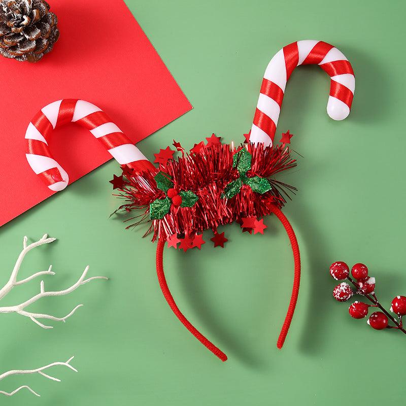Festive headband with red tinsel, green leaves, and candy cane antennas on a green background. Nearby are a pine cone, white twig, red berries, and a red card.