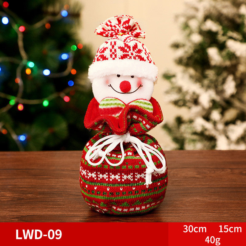 Festive snowman decoration with a red and green knit hat and scarf, smiling on a wooden table. Christmas tree with colorful lights blurry in the background.