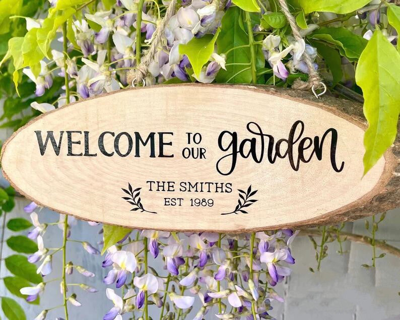 Wooden sign with "Welcome to our garden, The Smiths, Est 1989" in black script. Surrounded by lush green leaves and purple wisteria flowers.