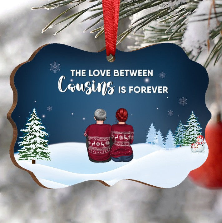 Ornament with two people in matching sweaters, snowy landscape, and trees. Text reads "The love between cousins is forever" with snowflakes and a snowman. Red ribbon attached.