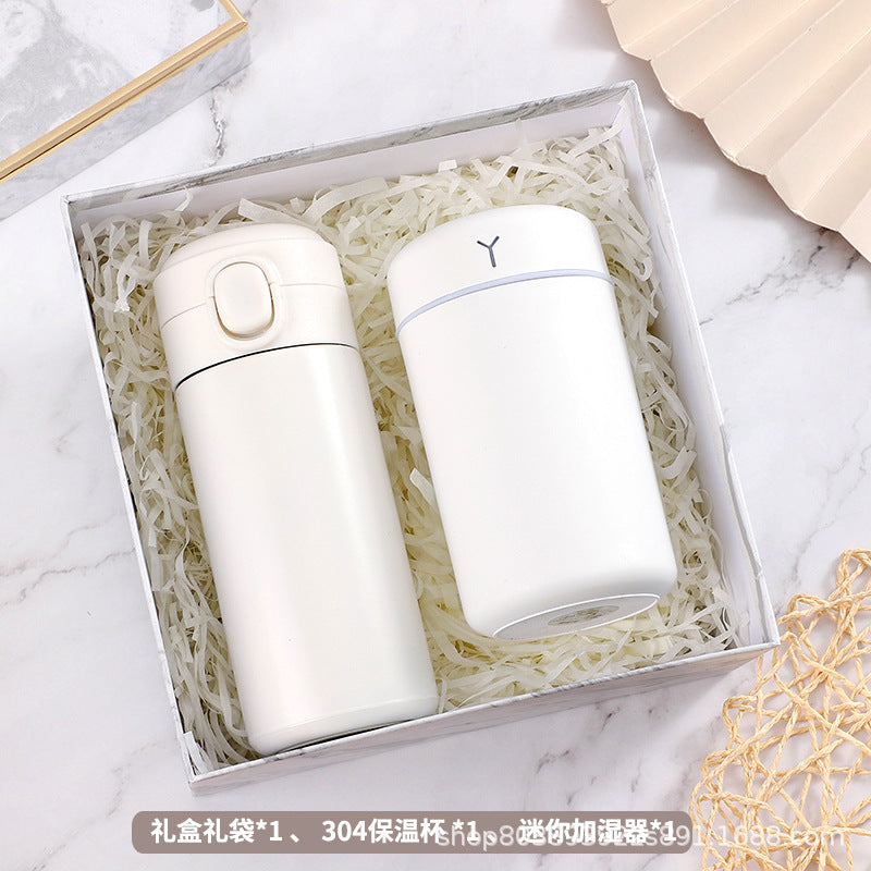 A gift box containing a white thermos and a white cylindrical device, nestled in shredded paper, placed on a marble surface.