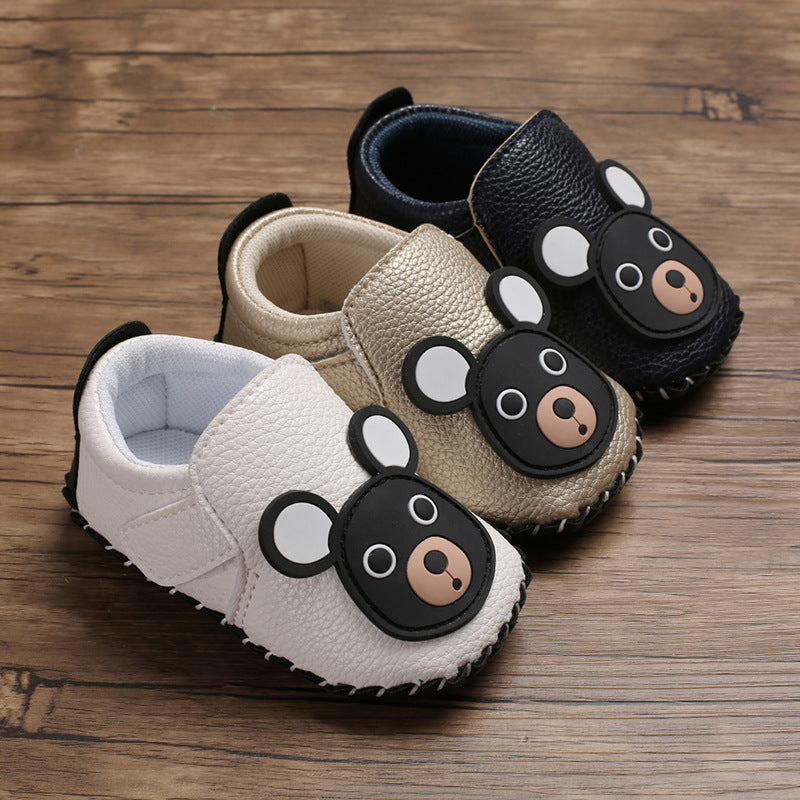 Three pairs of toddler shoes on wooden floor, featuring bear faces. Colors include white, beige, and black. Each shoe has a bear design with ears and a nose.